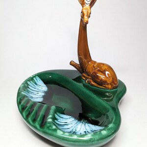 May include: A green ceramic ashtray with a brown deer figurine on top. The ashtray has two compartments and white accents.