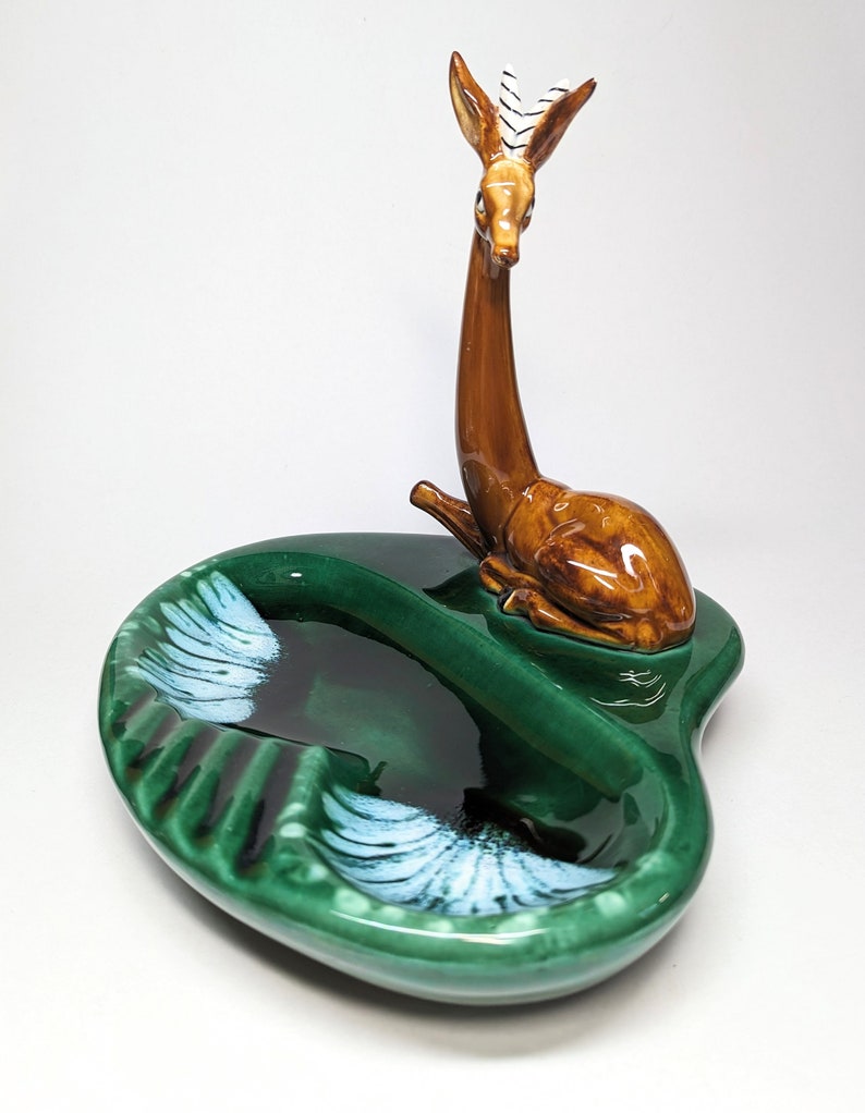 May include: A green ceramic ashtray with a brown deer figurine on top. The ashtray has two compartments and white accents.