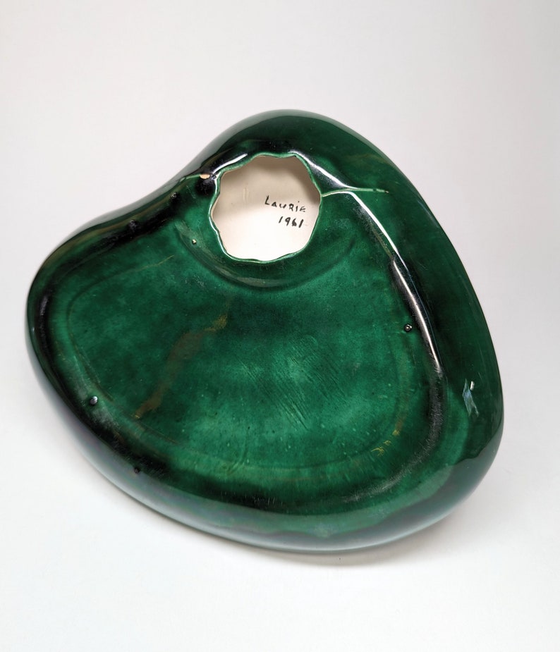 May include: A green ceramic heart-shaped dish with a hole in the center. The bottom of the dish is signed "Laurie 1961".