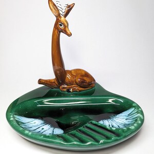 May include: A green ceramic ashtray with a deer figurine on top. The deer has brown fur, black eyes, and white stripes on its antlers. The ashtray has a heart shape with a divided section for cigarettes and a larger section for ashes.