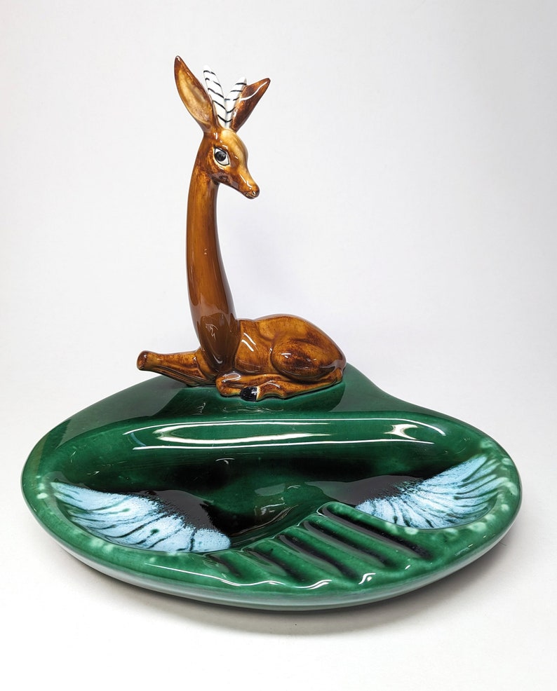 May include: A green ceramic ashtray with a deer figurine on top. The deer has brown fur, black eyes, and white stripes on its antlers. The ashtray has a heart shape with a divided section for cigarettes and a larger section for ashes.