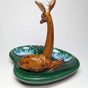 May include: A green ceramic ashtray with a brown deer figurine on top. The deer has white stripes on its horns and is looking to the left. The ashtray has a blue and white floral design.