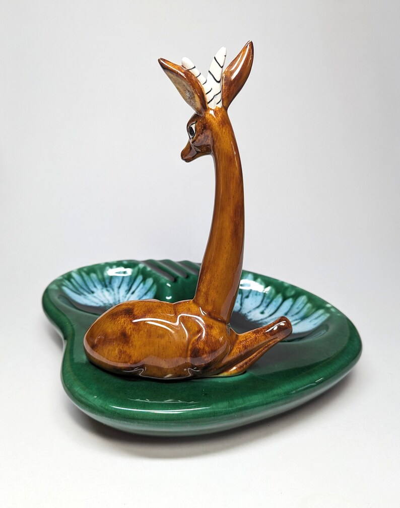 May include: A green ceramic ashtray with a brown deer figurine on top. The deer has white stripes on its horns and is looking to the left. The ashtray has a blue and white floral design.