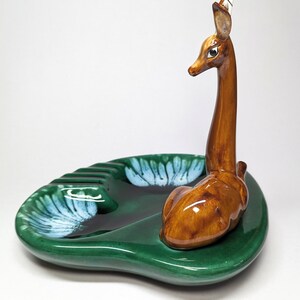 May include: A green ceramic ashtray with a deer figurine on top. The deer is brown with white accents and has a long neck. The ashtray has a leaf design and is shaped like a leaf.