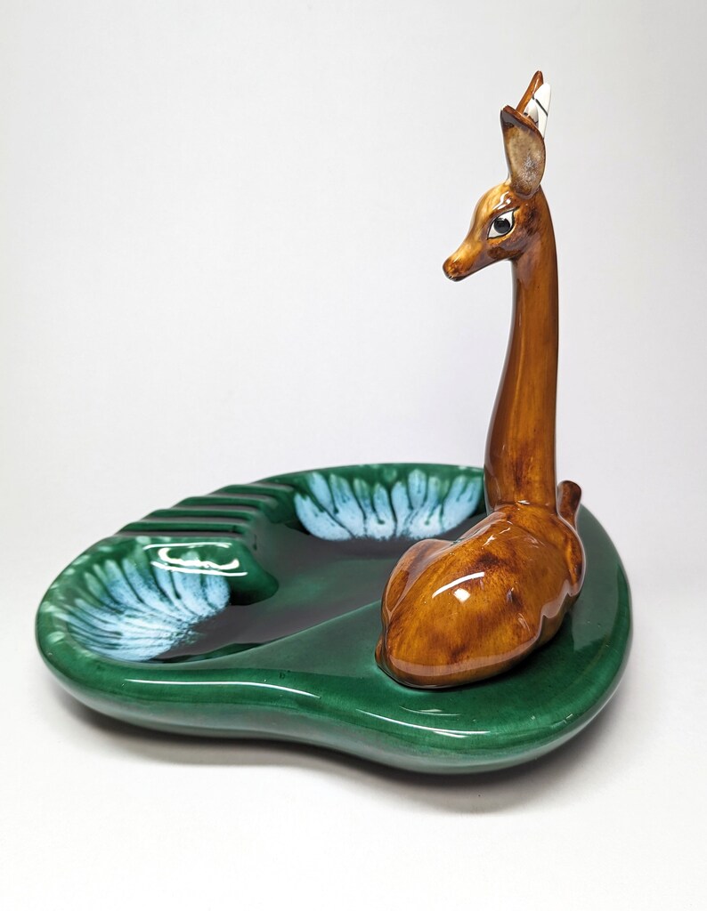 May include: A green ceramic ashtray with a deer figurine on top. The deer is brown with white accents and has a long neck. The ashtray has a leaf design and is shaped like a leaf.