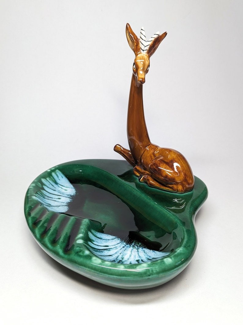 May include: A green ceramic ashtray with a deer figurine on top. The ashtray has two compartments and white accents.