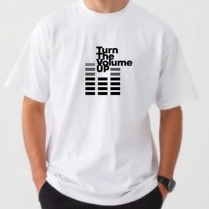 May include: White t-shirt with the text "Turn The Volume UP" in black and gray. The graphic includes a sound wave design. The shirt is short-sleeved and appears to be made of a soft material.