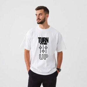 Pode incluir: Camiseta branca com o texto preto "TURN THE UP". O gráfico apresenta um design que se assemelha aos faders de mixagem de som. A camisa é um estilo casual e unissex.