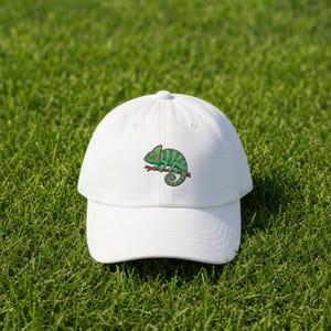 May include: A white baseball cap with an embroidered design of a green and teal chameleon on a brown branch. The cap is made of a soft material and has a curved peak. The cap is sitting on green grass.