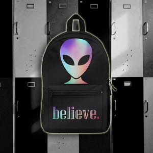 May include: Black backpack featuring a holographic alien head design and the word "believe" in a rainbow gradient. The backpack has a front pocket and a top handle, perfect for school or travel.