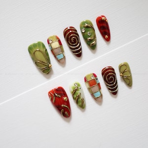 Op de afbeelding: Een set van tien kunstnagels met amandelvormige punten en gevarieerde ontwerpen. De nagels hebben een kleurenpalet van olijfgroen, donkerrood en crème, geaccentueerd met gouden metalen krullen, geometrische rasterpatronen en kleine strass-steentjes.