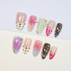 Puede incluir: Una colección de diez uñas artificiales con varios diseños. Algunas uñas presentan tachuelas doradas, flores rosas y patrones abstractos. Otras tienen una base nude con acentos rojos. Las uñas están dispuestas sobre una superficie blanca.