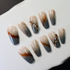 Peut inclure: Ensemble de dix faux ongles avec un design dégradé. Les ongles sont de couleur beige clair à la base et passent au gris foncé à la pointe. Certains ongles ont des accents dorés.