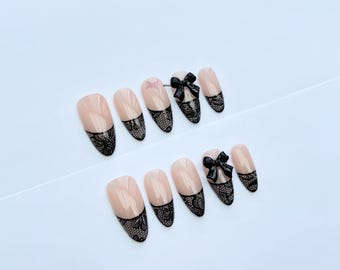 Può includere: Un set di unghie finte a forma di mandorla. Le unghie sono di colore nude con un design in pizzo nero sulle punte. Alcune unghie presentano un fiocco nero. Le unghie sono disposte su una superficie bianca.