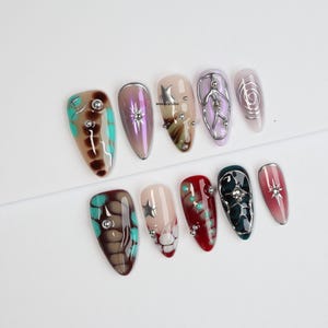 May include: A set of ten artificial nails with diverse designs. The nails display colours including brown, teal, purple, red, and pink, with silver studs, stars, and swirls. The nails are arranged on a white surface.