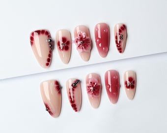 May include: A set of ten almond-shaped artificial nails. The nails feature a nude base with burgundy floral designs, 3D flower accents, and silver bead embellishments. The nails are arranged on a white surface.