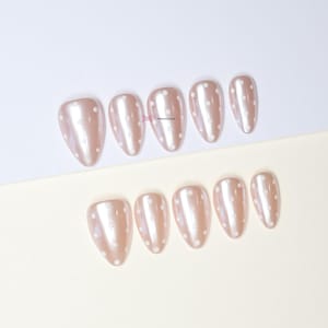 May include: A collection of almond-shaped false nails in a pearlescent finish. The nails are a light beige with a white polka dot design. They are arranged in two rows against a white and beige backdrop.