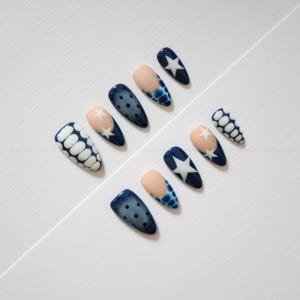 May include: A set of ten almond-shaped press-on nails featuring a variety of designs including dark blue with white stars, dark blue with black polka dots, nude tips with blue scale patterns, and white oval patterns on a dark blue base.