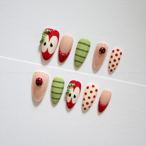 May include: A set of ten press-on nails featuring various designs including red apple shapes with green worms, green striped patterns, beige nails with ladybug accents and white scalloped edges, and cream nails with red polka dots or red tips.