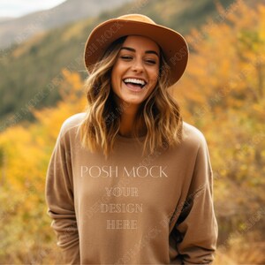 Puede incluir: Una mujer con una sudadera marrón con el texto "POSH MOCK YOUR DESIGN HERE" y un sombrero marrón, sonriendo en un campo de hojas amarillas y naranjas.