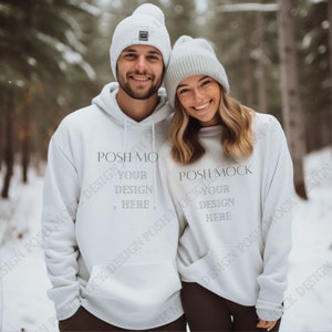 Puede incluir: Dos sudaderas con capucha blancas con el texto "POSH MOCK DESIGN YOUR DESIGN HERE" en ellas. La pareja lleva gorros blancos y están de pie en un bosque nevado.