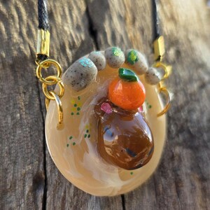 May include: A handcrafted pendant necklace with a brown capybara and an orange fruit. The pendant is set in a light-coloured resin with gold-coloured accents and a black cord. The pendant is approximately 3.8 cm in diameter.