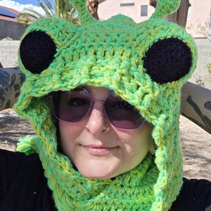 May include: A bright lime green crochet hood featuring two large, round black eyes and two small antennae on top. The hood is constructed with a thick, textured yarn weave and covers the head and neck area, providing a snug fit around the face.