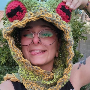 May include: A green, yellow, and gray crocheted frog hat with red and black eyes. The hat is worn by a person with glasses and a nose ring.