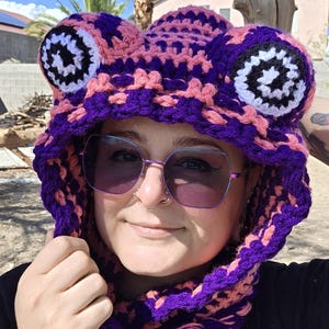 May include: A crochet hood featuring a frog design with large circular eyes in white, black, and purple. The hood is constructed from thick yarn in alternating stripes of vibrant purple and soft pink, covering the head and neck area completely.