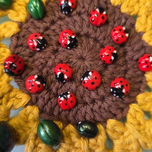 May include: A crocheted sunflower coaster in yellow and brown yarn, decorated with several small red ladybug-shaped beads and metallic green beetle-shaped beads scattered across the dark brown center of the flower-shaped textile piece.
