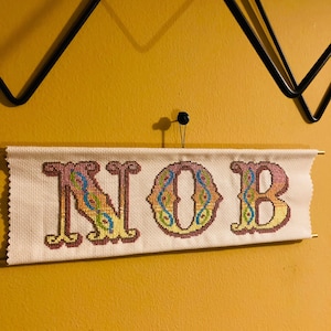 May include: A white cross-stitch with the word "NOB" in colorful, stylized lettering. The letters are outlined in red and filled with a gradient of yellow, orange, and pink. The background is a white fabric with a subtle grid pattern.