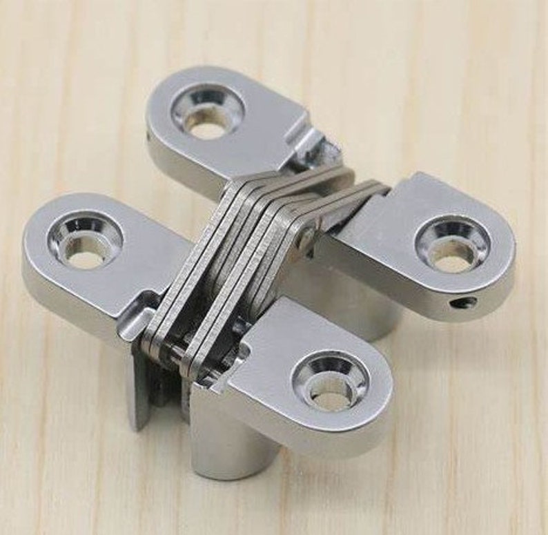May include: A polished, silver-colored concealed hinge. The hinge is designed with multiple interlocking plates and rounded mounting plates with screw holes. The hinge is designed for a flush, hidden installation.
