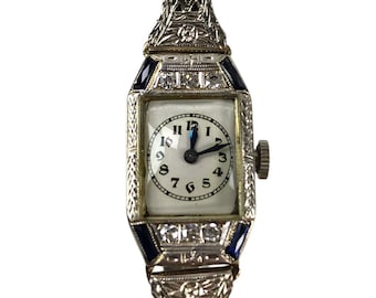 Könnte beinhalten: Eine Vintage-Armbanduhr mit weißem Zifferblatt und schwarzen römischen Ziffern. Die Uhr hat ein silberfarbenes Metallband mit geometrischem Muster und ist mit blauen Saphiren und Diamanten verziert.