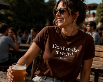 May include: A brown t-shirt with the text "Don't make it weird. please." in white. The person is holding a glass of beer. The image is taken outdoors on a sunny day.