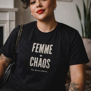 May include: Black t-shirt with the words "FEMME CHAOS" in white, with a star graphic. Below the text is "Non-Binary Collection". The person in the image has short, dark curly hair and a tattoo on their arm.