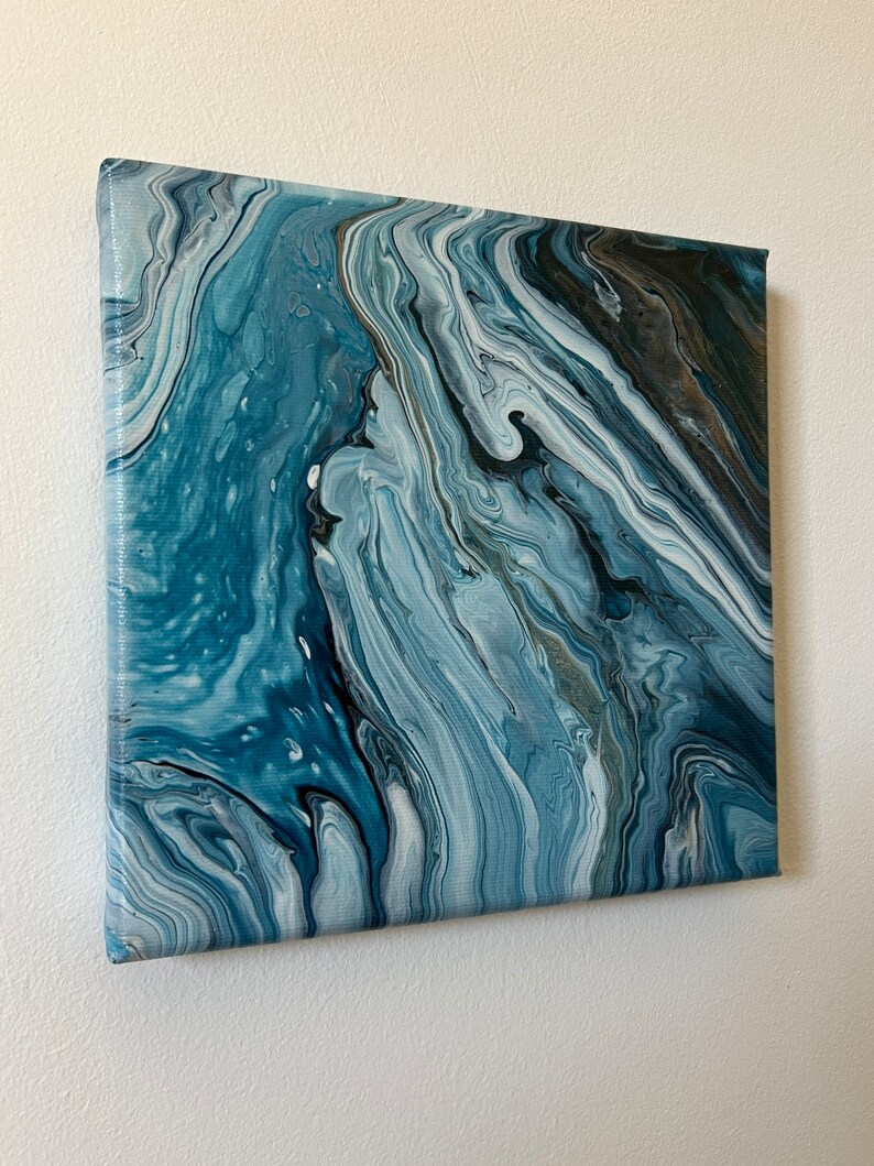 Puede incluir: Pintura abstracta sobre lienzo con colores azules, blancos y grises que se arremolinan. La pintura tiene un aspecto fluido y org&aacute;nico.