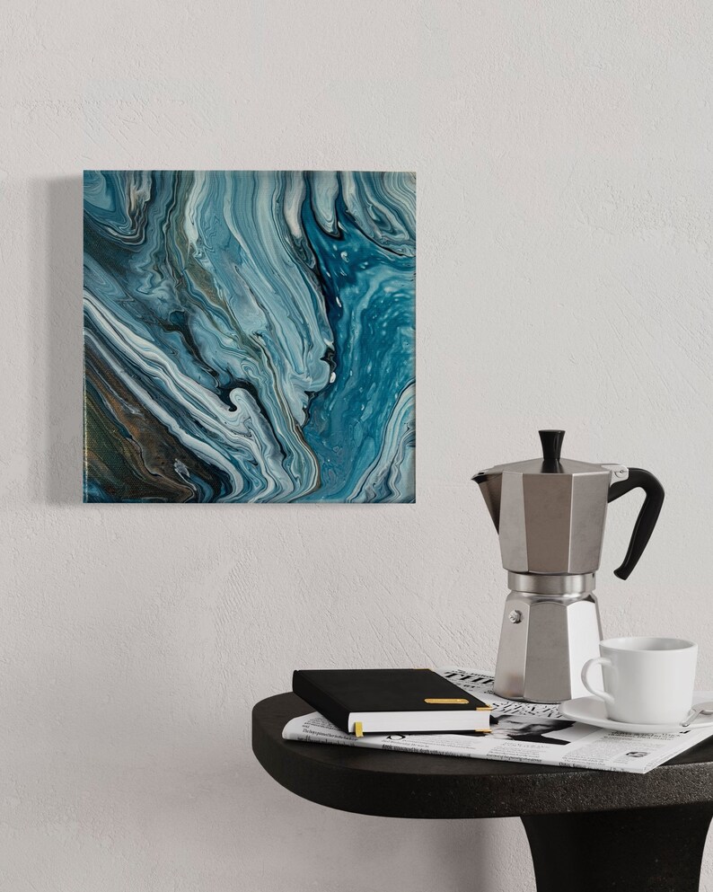 Puede incluir: Una pintura abstracta cuadrada con pintura azul, blanca y gris en espiral. La pintura est&aacute; colgada en una pared blanca sobre una mesa auxiliar negra con una cafetera, una taza y un platillo, y un cuaderno negro encima de un peri&oacute;dico.