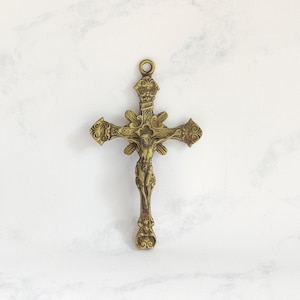 May include: An antique brass-toned metal crucifix pendant featuring an ornate, detailed design with decorative scrollwork at the ends of each arm and a central figure of Jesus Christ. A small circular loop is positioned at the top for hanging.