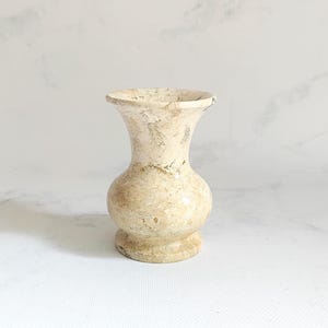 May include: A small, beige, natural stone vase with a porous, textured surface. The vase features a rounded, bulbous body, a narrow neck, and a flared, circular rim, resting on a sturdy, circular base with visible mineral patterns throughout.