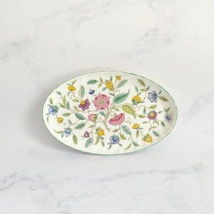 May include: An oval ceramic serving dish with a floral design. The dish has a cream base with a green rim, decorated with pink, yellow, blue, and green flowers and foliage. A decorative piece for serving food or displaying items.