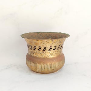 May include: A vintage brass planter with a scalloped rim and a decorative pierced design. The planter has a cylindrical shape with a ribbed base and a warm, aged patina. It is a decorative item for displaying plants or flowers.
