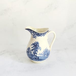 May include: Cream ceramic jug with a blue and white pastoral scene. The jug has a curved spout, a handle, and a detailed landscape design. The design includes trees, buildings, and figures, creating a classic, vintage aesthetic.