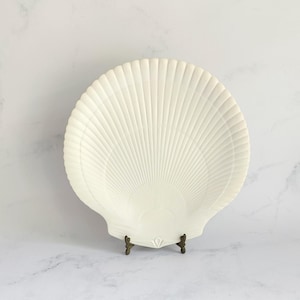 May include: A cream-coloured, shell-shaped decorative plate. The plate features a ribbed texture, radiating from the centre, mimicking a scallop shell. It is displayed on a small, bronze-coloured stand.