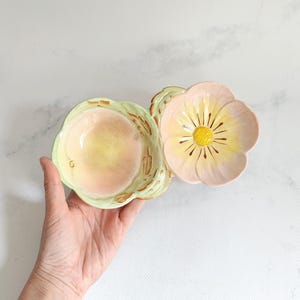 May include: A floral-shaped ceramic dish with a light pink and yellow color scheme. The dish has a flower-like design with a yellow center and pink petals. The dish is held by a hand.