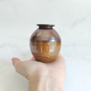 May include: A small, handcrafted brown pottery vase with a rounded body and a narrow neck. The vase has a glossy finish and a two-tone color scheme, with a darker brown top and a lighter brown base. The vase is held in a hand.