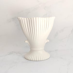 May include: A cream-coloured ceramic vase with a fluted design. The vase has a wide, flared top and a pedestal base. The exterior features vertical ridges, and small handles are on either side of the vase.
