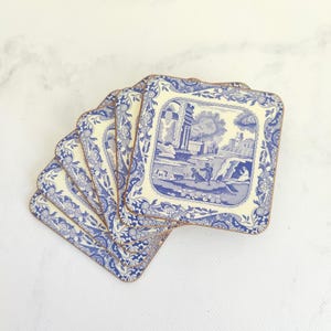 May include: A set of square coasters featuring a blue and white landscape design. Each coaster has a decorative border and a light cream background. The coasters are made from a durable material and are suitable for use with hot or cold drinks.