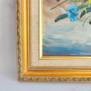 May include: Oil painting of a floral arrangement in a gold ornate frame. The artwork features blue, orange, and green flowers with a light blue and purple background. The frame is a warm gold color with detailed carvings.