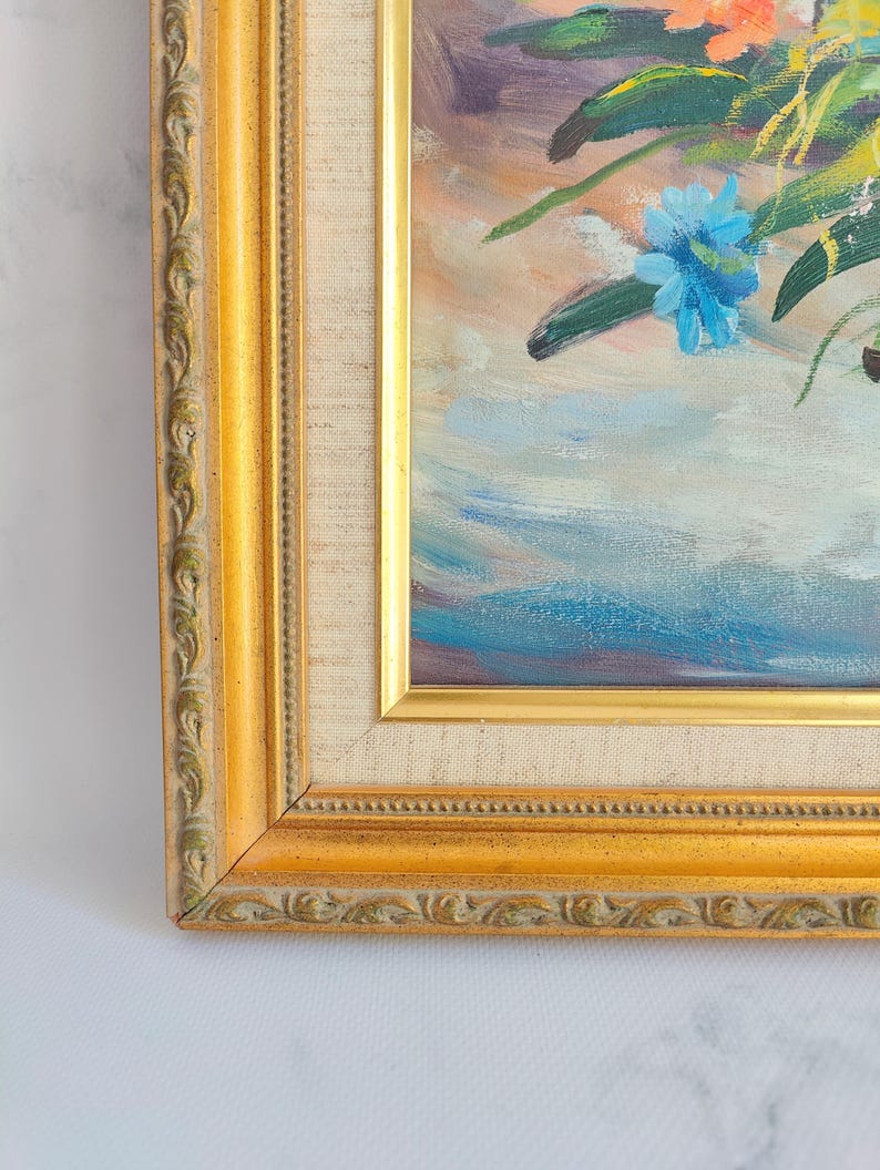 May include: Oil painting of a floral arrangement in a gold ornate frame. The artwork features blue, orange, and green flowers with a light blue and purple background. The frame is a warm gold color with detailed carvings.