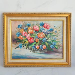 May include: Oil painting of a floral arrangement in a basket, framed in a gold-colored ornate frame. The flowers are a mix of pink, orange, and blue, with green leaves. The background is a soft, light color.